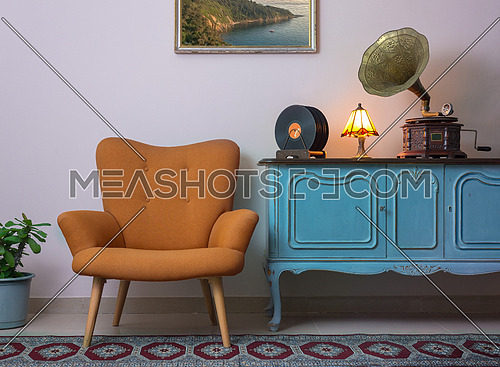 Vintage interior of retro orange armchair, vintage wooden light blue sideboard, old phonograph (gramophone), vinyl records and illuminated table lamp on background of beige wall, and red carpet