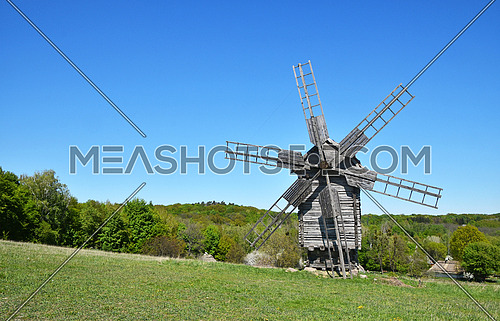 Rural meadow landscape with old wooden windmill, forest trees and clear blue sky