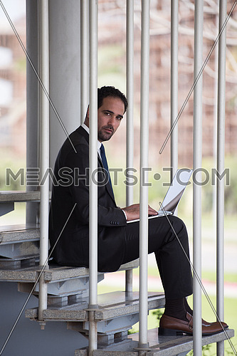 Young handsome middle eastern business executive sitting on steps in a corporate offices building using laptop