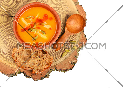 Small ceramic bowl of pumpkin cream soup, wooden spoon, slice of bread and seeds on wood cut isolated on white background, top view