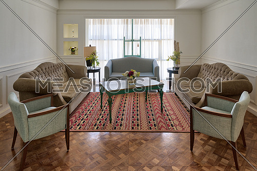 Interior shot of luxury modern living room with three couches, two armchairs, and vintage wooden table on background of big window with white curtains and decorated carpet on barquet floor