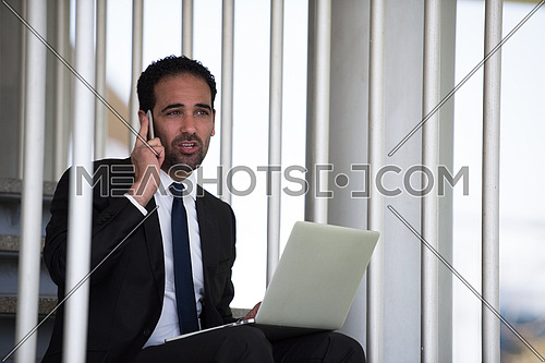 Young middle eastern business executive sitting on the stairs of a corporate office working with his laptop and talking on the phone
