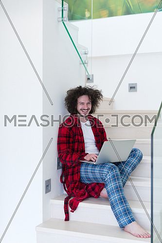 young freelancer in bathrobe working from home using laptop computer while sitting on stairs