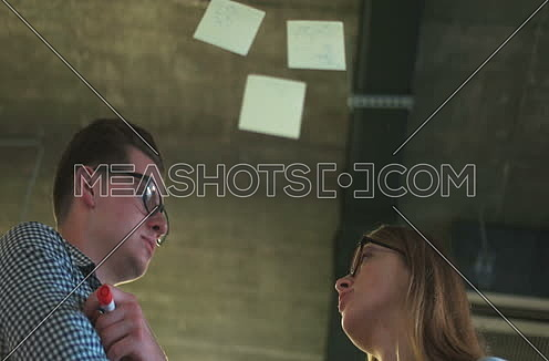Low angel for two business partners discussing sticky notes on glass wall at office interior.