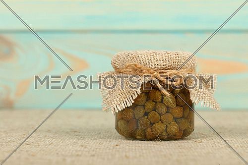 Close up of one small glass jar of pickled capers with canvas top decoration and twine on canvas tablecloth over blue wooden background, low angle side view