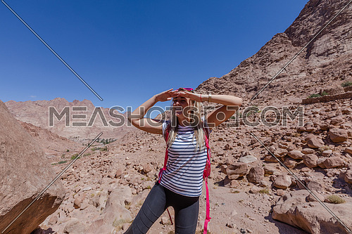 long shot for a female tourist wearing a pink Glasses and travel backpack climbing a big rock to explore Sinai Mountain for wadi Freij at day.