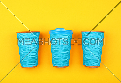 Close up three crimped disposable pastel blue paper takeaway coffee cups with plastic cap over vivid yellow background, flat lay, elevated top view, directly above