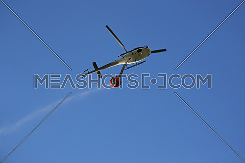 Firefighting helicopter with water bucket and trail of drops seen from below against blue sky 