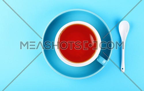 Close up one full cup of black or fruit red tea on saucer with white spoon over pastel blue paper background, elevated top view, directly above