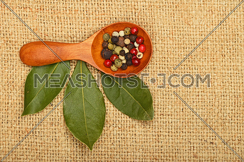 Three green bay laurel leaves and mix of peppercorn in wooden scoop on brown burlap jute canvas background