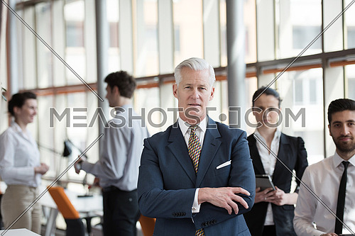 portrait of senior businessman as leader  at modern bright office interior, young  people group in background as team