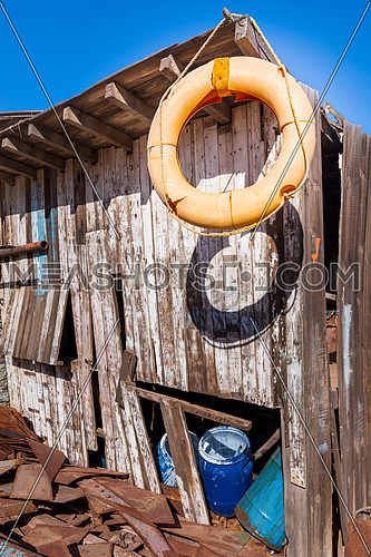 Old orange life saver hanged on edge of roof of shabby small wooden building among lot of boards and clear blue sky