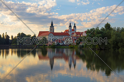 Our Lady Monastery (The Abbey of Nova Rise) with reflection over calm waters of lake and cloudy sunset sky in Telc, Moravia, Czech Republic