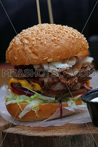 Big American burger with pulled pork meat, onion rings, cheese and salad in sesame bun on paper parchment over natural wooden cut, close up, low angle view
