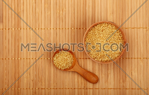 Wooden round scoop spoon and small bowl of brown cane sugar on bamboo mat background, top view