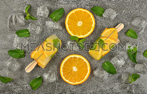 Close up two fruit ice cream popsicles with fresh orange slices, green mint leaves and ice cubes on gray table surface, elevated top view, directly above