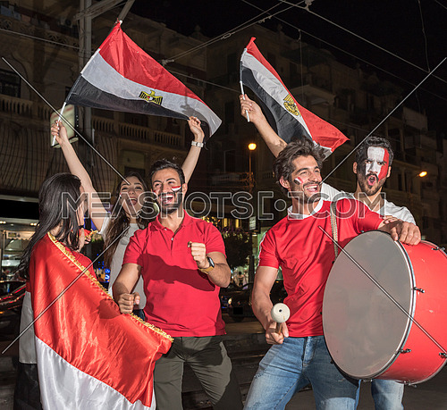 group of young people cheering for egypt, holding egyptian flags and drum while one of them has a paint egyptian flag on his face in the korba area at night
