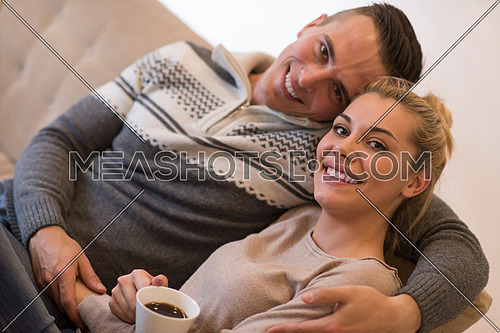 Young romantic couple sitting on sofa in front of fireplace at home, looking at each other, talking and drinking coffee autumn day