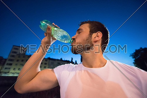 young athletic man drinking water after a night running session in the city