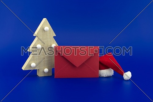 Red envelope for greetings next to a Christmas tree and Santa hat on a festive blue background. New Year and Christmas greeting season concept