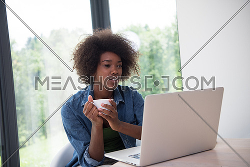 Young african american woman smiling sitting near bright window while looking at open laptop computer on table and holding white mug in her luxury home