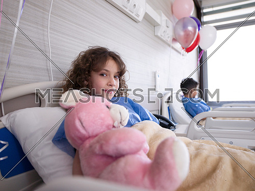 young girl lies in a hospital bed while hugs pink teddy bear