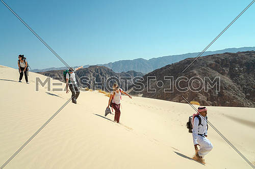 Long shot for group of tourists walking on sands with bedouin guide while exploring Sinai Trail from Ain Hodouda by day.