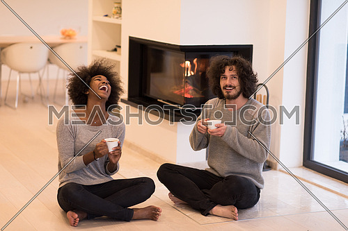 Young romantic multiethnic couple sitting on the floor in front of fireplace at home, looking at each other, talking and drinking coffee at autumn day