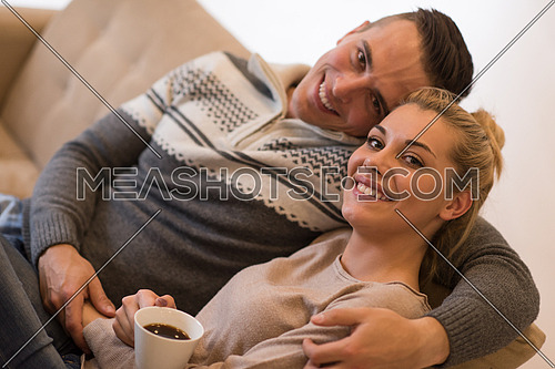 Young romantic couple sitting on sofa in front of fireplace at home, looking at each other, talking and drinking coffee autumn day