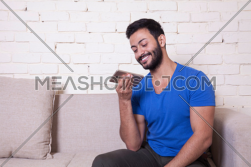 A young man sitting on a sofa using his Mobile Phone behind a white wooden table