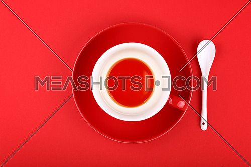 Empty cup of black tea on porcelain saucer with white ceramic spoon over vivid red paper background, close up, elevated top view, directly above