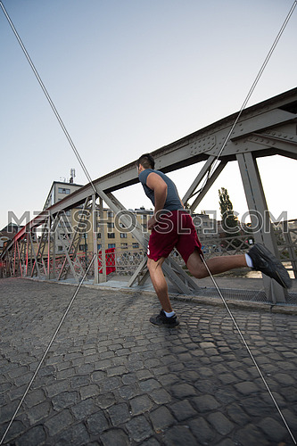 young sporty man jogging across the bridge at sunny morning in the city