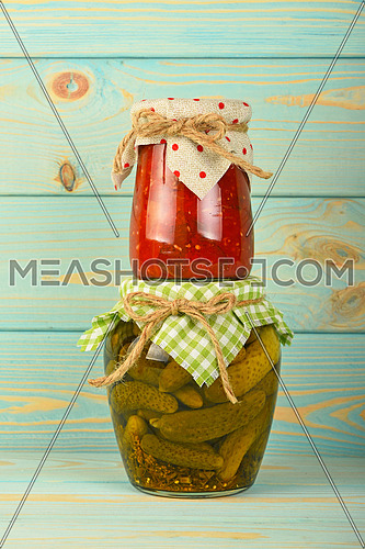 One glass jar of homemade eggplant pepper salad on jar of pickled cucumbers with green checkered textile decoration over blue painted wooden surface