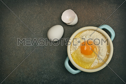 Close up flat lay of baking ingredients on dark grunge stone table surface with copy space, flour and eggs in pannikin, elevated top view, directly above