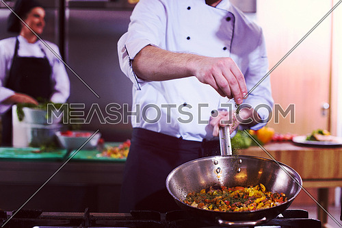 Young male chef putting spices on vegetables in wok at commercial kitchen