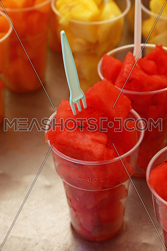 Street food, selection of fruit salads, slices of cut fresh ripe watermelon and melon cubes in plastic cups with forks at retail market stall display, close up, high angle view