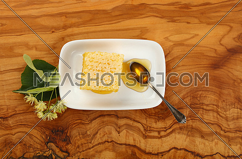 Close up fresh acacia honey with honeycomb, spoon and linden flowers on white plate over wooden table, elevated top view, directly above