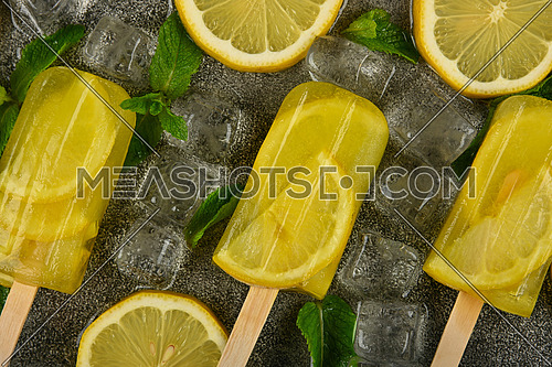 Close up frozen fruit juice popsicles with fresh lemon slices, green mint leaves and ice cubes on gray table surface, elevated top view, directly above