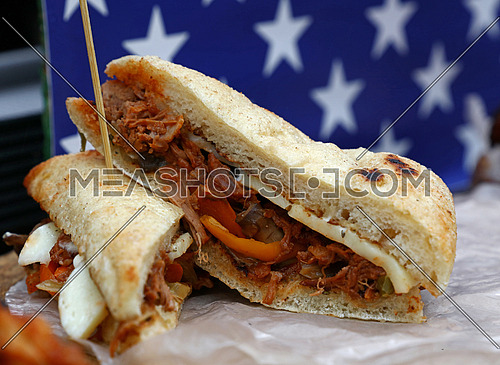 Two halves of one fresh grilled sandwich with pulled pork and cheese on paper parchment under American flag, close up, low angle view