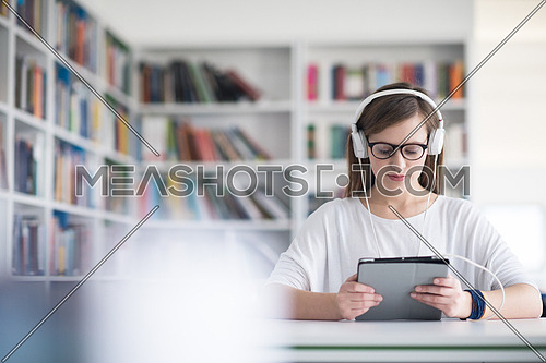 female student study in school library, using tablet and searching for informationâs on internet. Listening music and lessons on white headphones