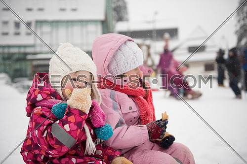 portrait of two cute little girls sitting together on sledges outdoors at snowy winter day, eating tasty cookies on break