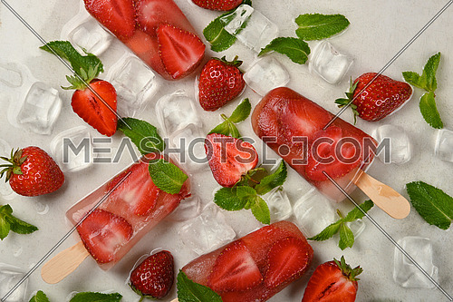 Close up fruit ice cream popsicle with fresh strawberry, green mint leaves and ice cubes on white table surface, elevated top view, directly above