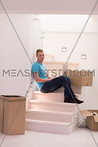 Happy young man sitting in stairway at home, using laptop computer with boxes around him