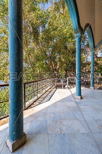 Elements of covered terrace with blue stone pillars and marble floor fenced with metal forged fence and surrounded with green trees in sunny summer day
