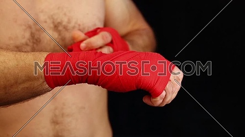 Close up man boxer tightening and testing red hand wraps over wrists preparing for fight, over black background, low angle front view