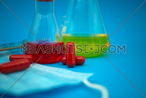 Medical, healthcare or Covid-19 still life with red capsules, hypodermic syringe, mask and chemical solution in a flask over a blue background
