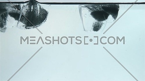 Clear ice cubes float and melt with bubbles of air in blue transparent water, extreme close up, low angle side view, slow motion