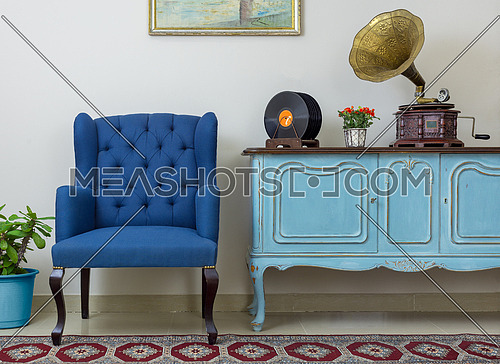 Vintage interior of retro blue armchair, vintage wooden light blue sideboard, old phonograph (gramophone), vinyl records on background of beige wall, tiled porcelain floor, and red carpet