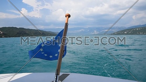 Close up flag of EU, European Union at back of boat, waving and blowing in the wind over blue sky and lake water, low angle view of action camera
