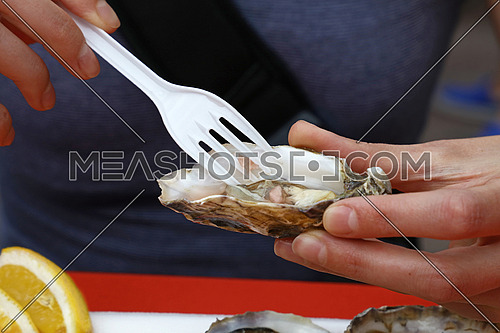 Young woman eating fresh raw open oyster with plastic fork at street seafood cafe, close up, low angle front view